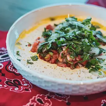 Creamy hummus topped with chopped tomatoes, onions, and fresh parsley, served in a white bowl at Feta Cowboy.